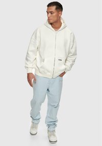 Hvid hættetrøje med lynlås og stor hætte, med kenguruposer. Bæres med lyseblå jeans og hvide sneakers med gule accenter.