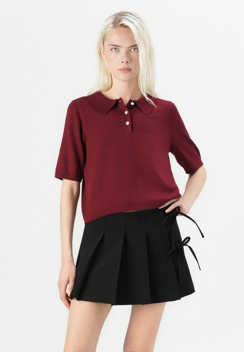 Blondynka ubrana w burgundowy sweterek polo z krótkim rękawem, zapinany na perłowe guziki, oraz czarną plisowaną mini spódnicę z bocznymi kokardkami.