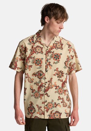 Giovane uomo che indossa una camicia beige a maniche corte con motivo paisley floreale rosso e verde, guardando in basso a sinistra.