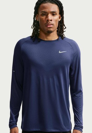 Uomo con dreadlocks fino alle spalle che indossa una maglia sportiva Nike a maniche lunghe blu navy su uno sfondo semplice.