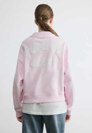 Person mit zurückgebundenem Haar, trägt ein pinkfarbenes Sweatshirt mit dem Schriftzug "Tommy Jeans" auf dem Rücken, ein weißes Hemd und blaue Jeans, blickt weg.