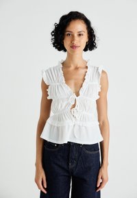 White peplum blouse with textured fabric, ruffled edges, deep V-neck, tie-front detail, and short sleeves, paired with dark denim.