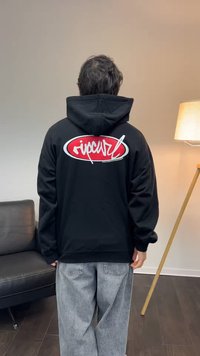 Persoon draagt een zwarte hoodie met rood-wit "ripcurl" logo op de achterkant, grijze spijkerbroek, staat binnen bij een zwarte bank en een vloerlamp.