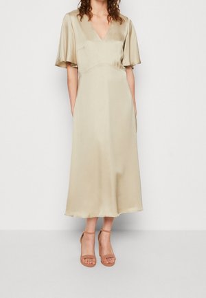 Vrouw die een beige midi-jurk van satijn met een V-hals draagt, korte fladdermouwen en sandalen met een enkelbandje en hoge hak in leerkleur.