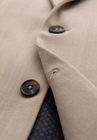 Gros plan d'un blazer en tissu chevrons beige avec des boutons marron foncé et un aperçu de la doublure intérieure à motif sombre.
