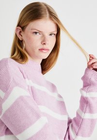 Lys lilla strikket genser med hvite striper, med høy krage og ribbede mansjetter, vist på en modell med langt, rett hår.