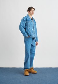 Τζάκετ από τζιν και τζιν παντελόνι σε ανοιχτό μπλε, με κουμπιά, πλαϊνές τσέπες και αντίθεση ραφής. Καφέ μπότες με κορδόνια ολοκληρώνουν το ντύσιμο.