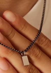 Zilveren rechthoekige hanger aan een zwarte ketting, met een sluiting en getextureerde schakels. Handen die de ketting voorzichtig vasthouden.