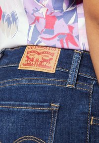 Jeans in denim blu scuro con un'etichetta in pelle con un motivo rosso, che mostra dettagli come rivetti e cuciture. Vista ravvicinata della tasca posteriore.