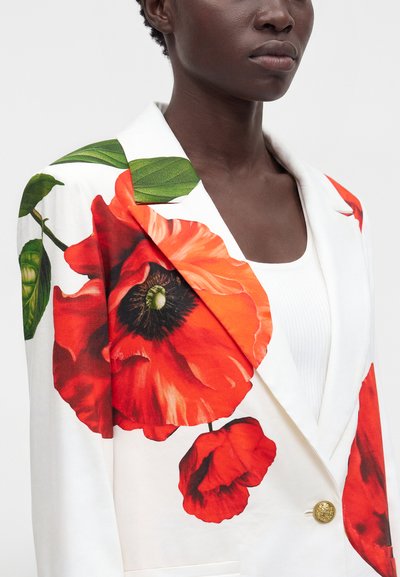 Personne noire portant un blazer blanc avec de grandes impressions de fleurs de coquelicot rouges et un bouton doré sur un haut blanc.