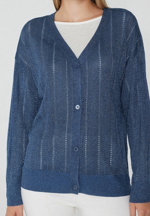 Persoon die een blauwe, dichtgeknoopte, lange mouwen cardigan met subtiele verticale strepen over een wit shirt draagt, gecombineerd met een witte broek.