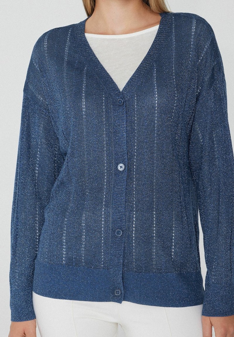 Personne portant un cardigan bleu boutonné à manches longues avec de fines rayures verticales sur un haut blanc, assorti à un pantalon blanc.
