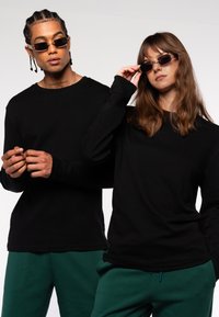 Schwarze Langarmshirts kombiniert mit grünen Jogginghosen. Der Mann trägt eine Sonnenbrille mit dünnen Rahmen; die Frau hat eine Sonnenbrille mit breiteren Rahmen.