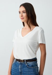 T-shirt bianco a V in tessuto morbido, con maniche corte. Abbinato a jeans in denim blu scuro e una cintura marrone con fibbia dorata.