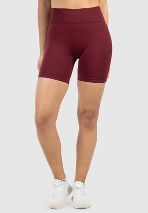 Een persoon staat in rode sportshorts en witte sneakers tegen een lichtgrijze achtergrond, wat de kledingstijl benadrukt.