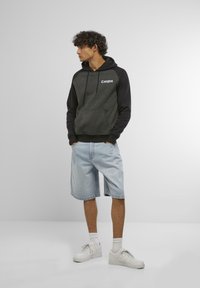 Sweatshirt à capuche en gris foncé et noir, avec le logo "Compton". Associé à un short en denim bleu clair et des baskets blanches.