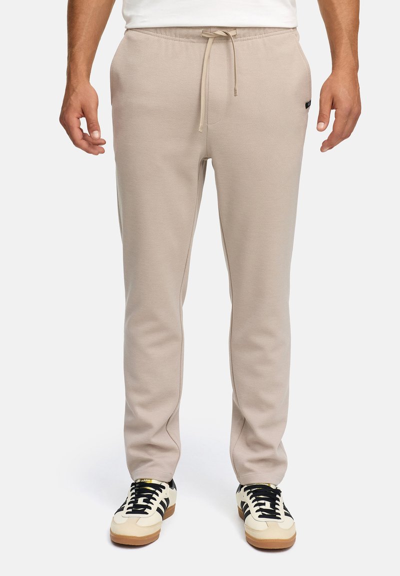 Man wearing beige jogger pants with drawstring waist and white sneakers with black stripes, standing against a white background.