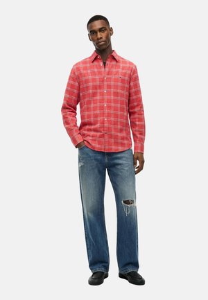 Mann trägt rotes kariertes Langarmhemd mit blauen zerrissenen Jeans und schwarzen Schuhen, steht mit einer Hand in der Tasche vor weißem Hintergrund.