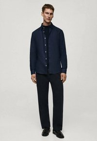 Camicia blu navy con bottoni sopra un dolcevita nero, abbinata a pantaloni scuri. Il modello indossa scarpe in pelle nere. Design minimalista e vestibilità su misura.