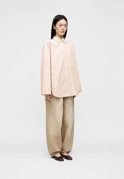 Femme aux cheveux noirs et raides portant une chemise beige rayée oversize à boutons, un pantalon beige large et des ballerines marron foncé, debout devant un fond gris.