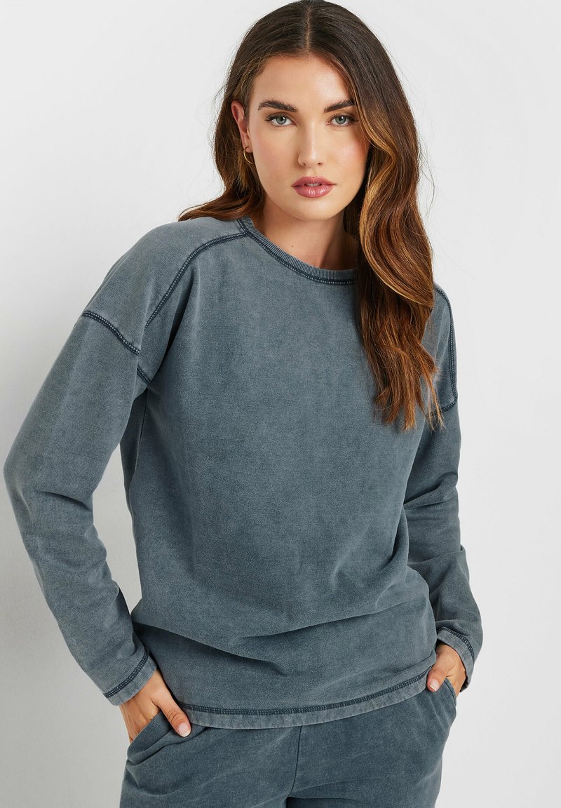 Grå sweatshirt i mjukt material med rund halsringning, långa ärmar och en avslappnad passform. Den har fickor och kontrasterande sömmar.