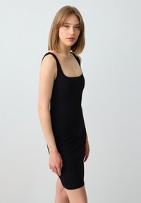 Schwarzes, tailliertes Kleid mit quadratischem Ausschnitt und breiten Trägern. Der Stoff ist glatt und dehnbar, was die Silhouette betont.