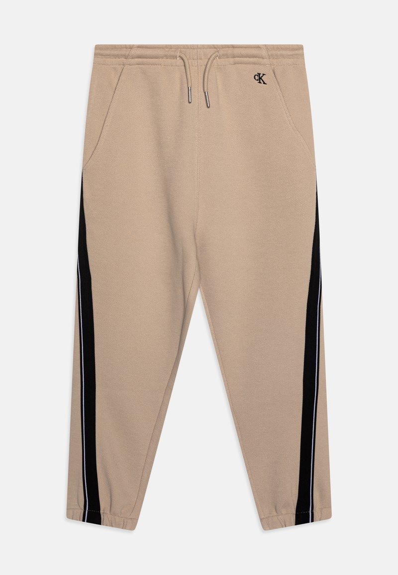 Beige Jogginghosen mit elastischem Bund, Kordelzug, Seitentaschen, schwarzen vertikalen Streifen an den Beinen und kleinem schwarzem "cK"-Logo auf der rechten Hüfte.