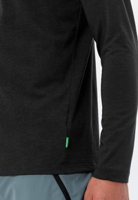 Maglietta nera a maniche lunghe realizzata in tessuto morbido, con un'etichetta verde sul lato inferiore, dal design aderente e dalla texture liscia.