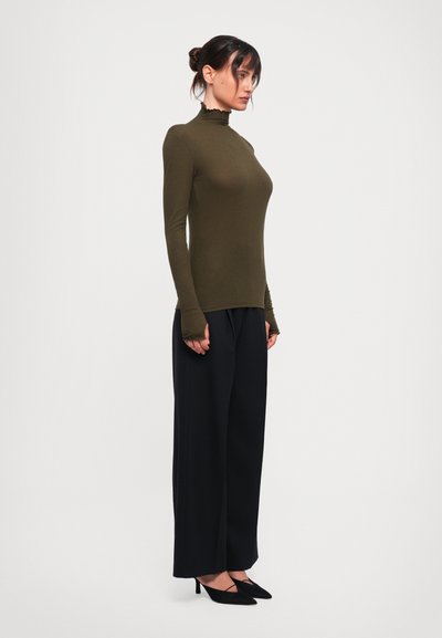Olive green long-sleeve top with a high neck and ruffled hem, paired with high-waisted black wide-leg trousers and pointed black heels.