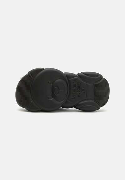 Suela de zapato de goma negra con forma de oso, que presenta detalles texturizados y un texto en relieve que dice "ESTO NO ES UN JUGUETE MOSCHINO."