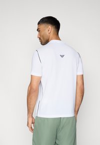 Λευκό πόλο με κοντά μανίκια και υφή, με μαύρες πλευρικές λεπτομέρειες και λογότυπο στο πάνω μέρος της πλάτης. Συνδυάζεται με πράσινα σορτς.