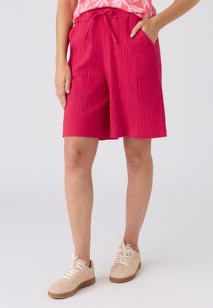 Vrouw draagt felroze gestructureerde knielange shorts met trekkoord in de taille, gecombineerd met beige sneakers op een lichte achtergrond.