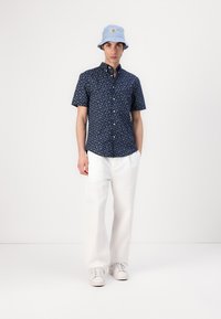 Chemise à manches courtes bleu marine à boutons avec un petit motif floral, associée à un pantalon blanc ample et un chapeau de pêcheur bleu clair. Des baskets blanches complètent le look.