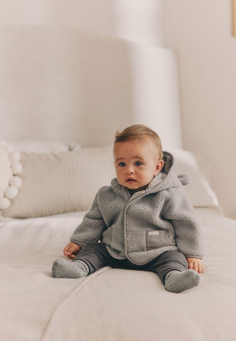 Graue Fleecejacke mit Kapuze, Frontreißverschluss und zwei Seitentaschen, getragen über grauen gestreiften Hosen und Socken, sitzt auf einem Bett.