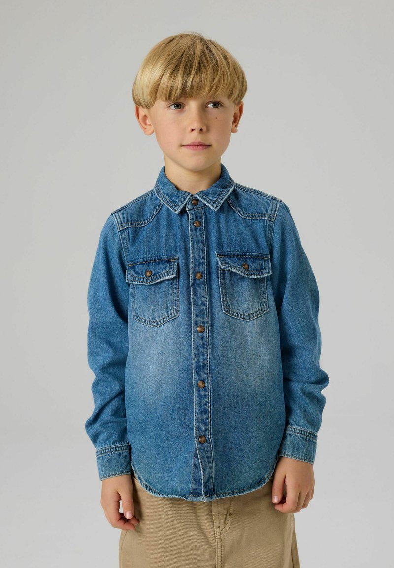 Vieglā zilā džinsa krekls ar pogām priekšpusē, asim formu apkakli, divām krūšu kabatām un garām piedurknēm. Mīksta tekstūra un izbalējis tonis.