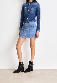 Giacca di jeans blu scuro con chiusura a bottoni e due tasche sul petto; abbinata a una minigonna di jeans blu chiaro con dettagli in cintura e tasche. Stivaletti neri alla caviglia.