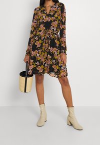 Schwarzes Blumenkleid mit langen Ärmeln, V-Ausschnitt, tailliertem Bund und lockerem Schnitt. Das Kleid hat pinke und gelbe Blumenmuster. Kombiniert mit beigen Stiefeln mit Absatz und einer Strohtasche.