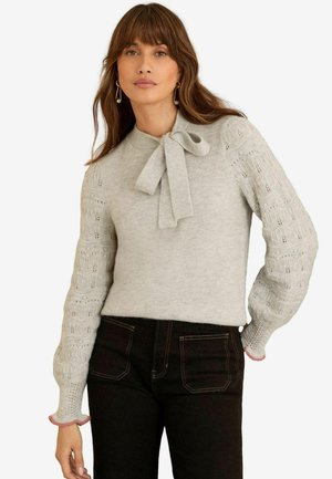 Hellgrauer Pullover mit Schleifenkragen, kompliziertem Muster, gestrickten Ärmeln und fransigen pinken Kanten. Kombiniert mit dunklen Jeans.