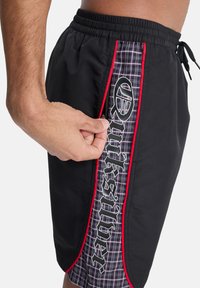 Μαύρο μαγιό με κόκκινες και καρό πλαϊνές λεπτομέρειες, με ελαστική μέση και λευκή λεπτομέρεια λογότυπου. Το χέρι προσαρμόζει το φερμουάρ.