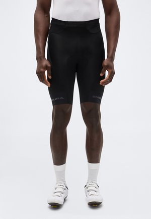 Férfi fekete kerékpáros rövidnadrágban, fehér zokniban és fehér kerékpáros cipőben, világos, egyszínű háttér előtt állva.