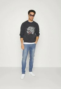 Νέος άνδρας με σκούρο φούτερ με γραφικό λιονταριού, μπλε τζιν, λευκά αθλητικά παπούτσια και γυαλιά ηλίου, στέκεται μπροστά σε απλή αχνή επιφάνεια.