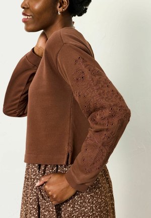 Braunes Sweatshirt mit bestickten Details an den Ärmeln, kurzen Ärmeln und Seitenschlitzen. Kombiniert mit einem braunen Rock mit Blumenmuster.