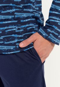 Camicia a maniche lunghe a righe blu con base navy; motivi in azzurro chiaro e verde teal; indossata con pantaloni blu scuro; mano appoggiata in tasca.