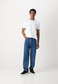 Camiseta blanca de algodón con un pequeño logo en el lado izquierdo del pecho, combinada con unos pantalones vaqueros de denim azul holgados y zapatos negros con suela blanca.
