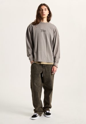 Převelký šedý sweatshirt s dlouhým rukávem a vyšitým textem, v kombinaci s volnými tmavě zelenými kalhotami a černými teniskami s bílými akcenty.