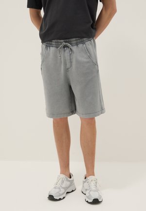 Mann trägt graue Shorts mit Kordelzug, schwarzes T-Shirt und weiße Sportschuhe, steht mit den Händen hinter dem Rücken vor einfarbigem Hintergrund.