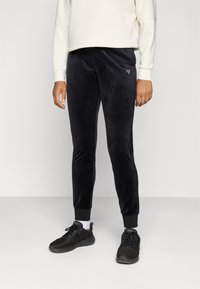 Zwarte velours joggers met taps toelopende pijpen en ribgebreide boorden, voorzien van een klein logo op de heup. Gepaard met zwarte sport schoenen.