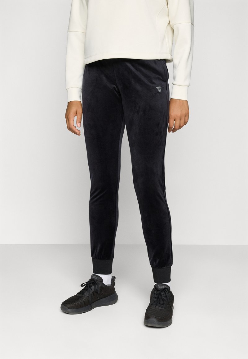Zwarte velours joggers met taps toelopende pijpen en ribgebreide boorden, voorzien van een klein logo op de heup. Gepaard met zwarte sport schoenen.