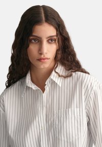 Biała koszula z długim rękawem w niebieskie pionowe paski, z kieszonką na piersi i klasycznym kołnierzykiem. Materiał wydaje się gładki i lekki.