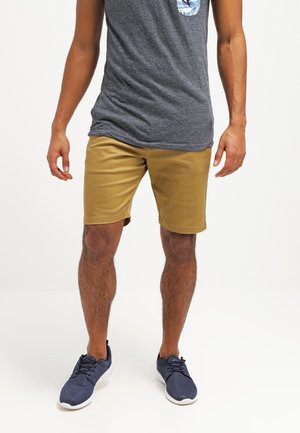 Mann trägt senfgelbe Shorts, marineblaue Turnschuhe und ein graues ärmelloses Hemd mit einem gemusterten Taschenaufnäher.